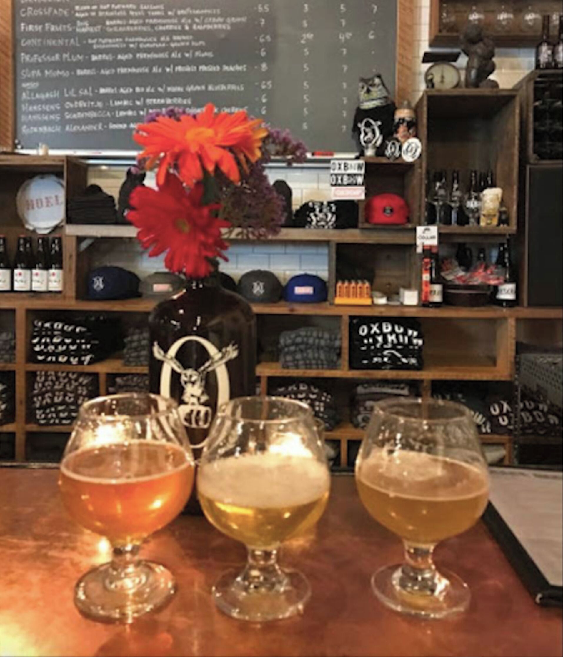 A bar area with three glasses of beer, a decorative vase with flowers, and shelves stocked with bottles and hats.
