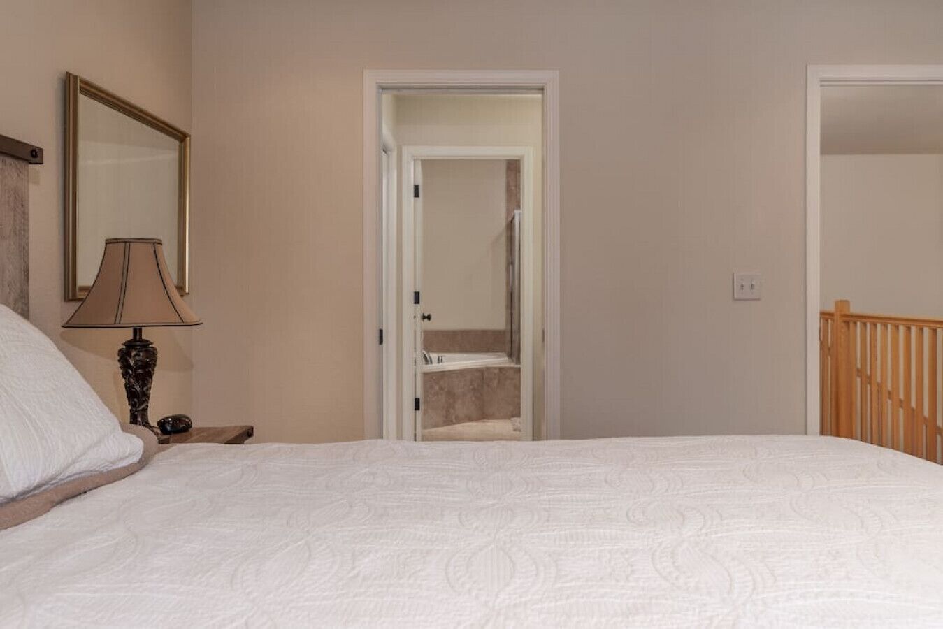 A bedroom with a bed, a nightstand, a lamp, a mirror, and a door leading to a bathroom.