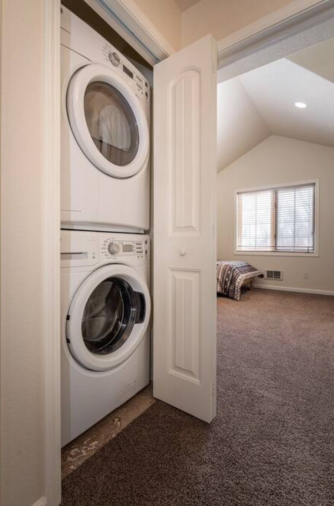 Uma lavanderia com duas máquinas de lavar e secar empilhadas, uma porta que dá para um quarto com janela e cama.