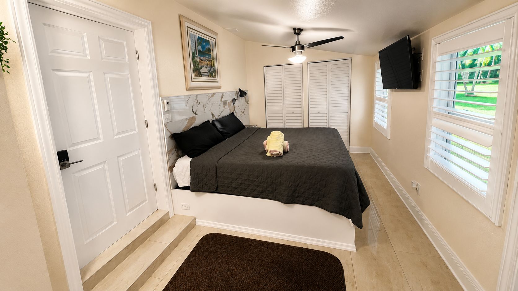 A bedroom with a bed, a ceiling fan, a TV mounted on the wall, and a window with blinds.