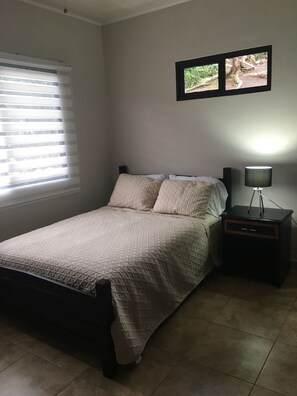 Un dormitorio con cama, mesita de noche, lámpara, ventana con persianas y un cuadro colgado en la pared.