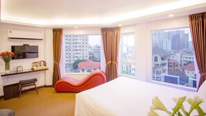 A hotel room with a bed, a desk with a flat-screen TV, a chair, a window with curtains, and a view of buildings. - Dinh Hotel (Hanoi)