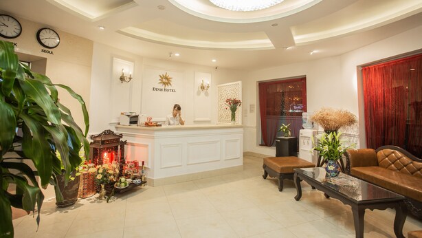A hotel reception area with a receptionist, a reception desk, a chandelier, a clock, and a large plant. - Dinh Hotel (Hanoi)