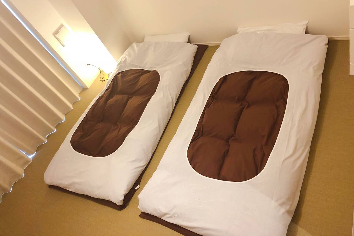 Two single beds with brown headboards and white bedding, positioned parallel to each other in a room with a light-colored carpet and a window with blinds.