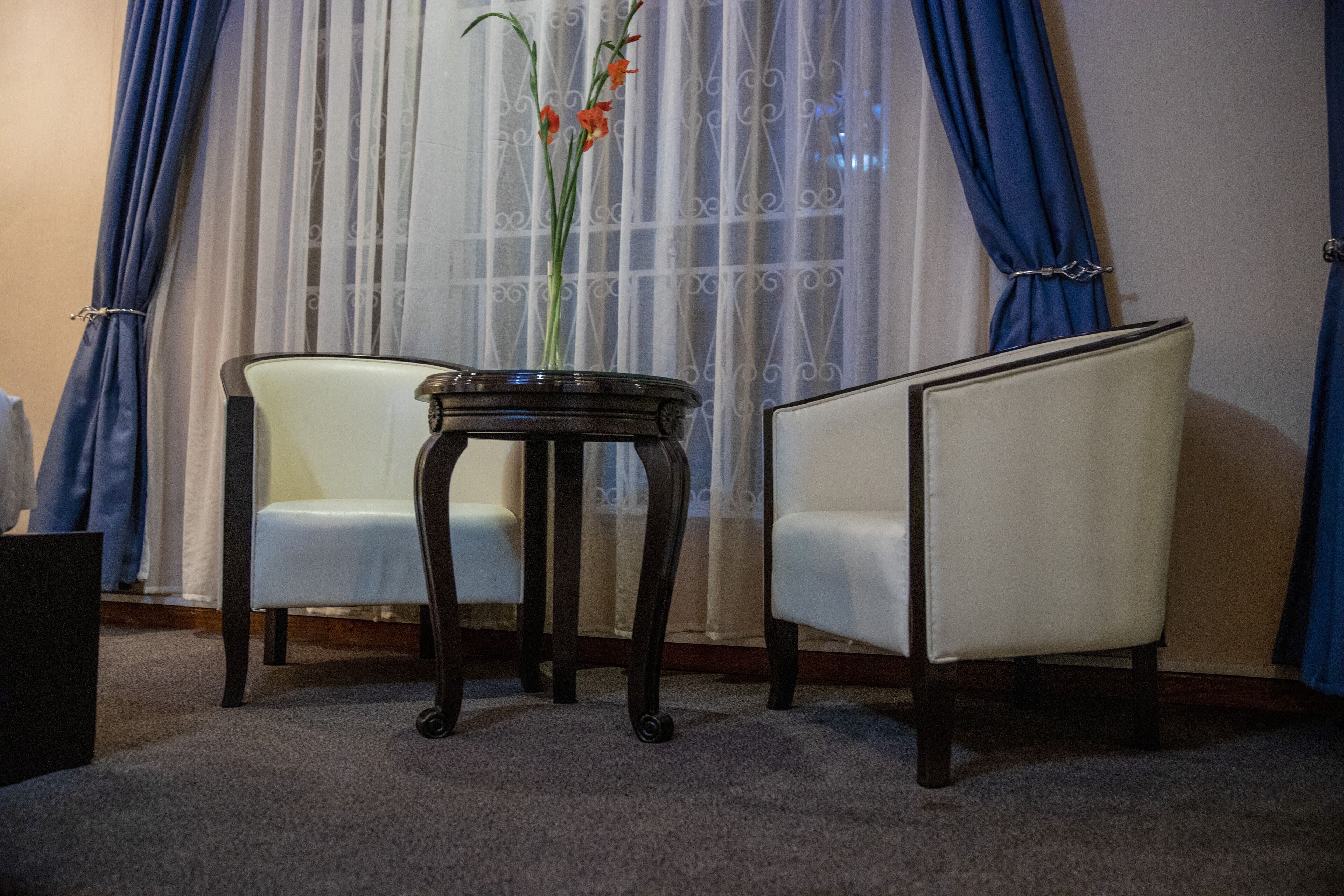 A room with two white upholstered chairs, a small round table with a vase of flowers, and a window with blue curtains.