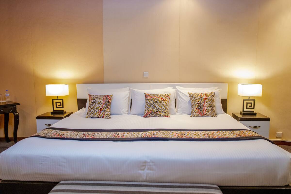 Una habitación de hotel con una cama grande, dos mesitas de noche con lámparas y una pared lisa.