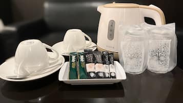 A table with a white electric kettle, two white cups, a plate with a spoon, a tray with six packets of cream powder, and two bags of hand sanitizer.