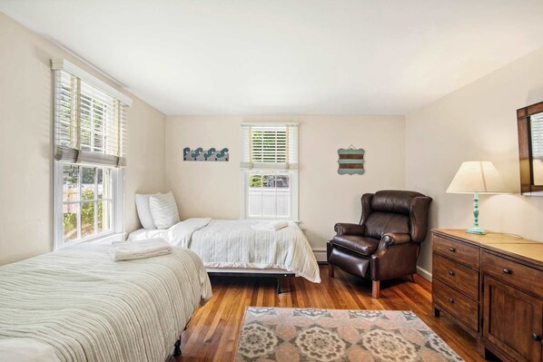 A bedroom with two beds, a leather armchair, a wooden dresser, and a window with blinds.