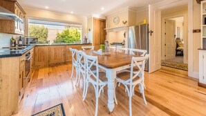 Une cuisine avec des meubles en bois, une table à manger entourée de chaises blanches, un réfrigérateur en acier inoxydable et une fenêtre donnant sur un jardin.