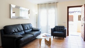 Ein Wohnzimmer mit schwarzem Ledersofa, einem Glastisch, einem schwarzen Sessel, einem großen Spiegel und Ausblick durch ein Fenster mit Vorhängen.