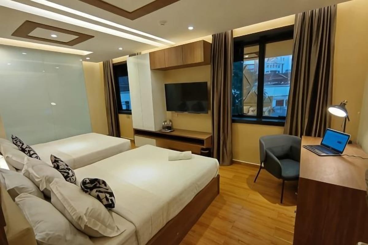 A hotel room with two beds, a desk with a laptop, a TV, and a window with curtains.