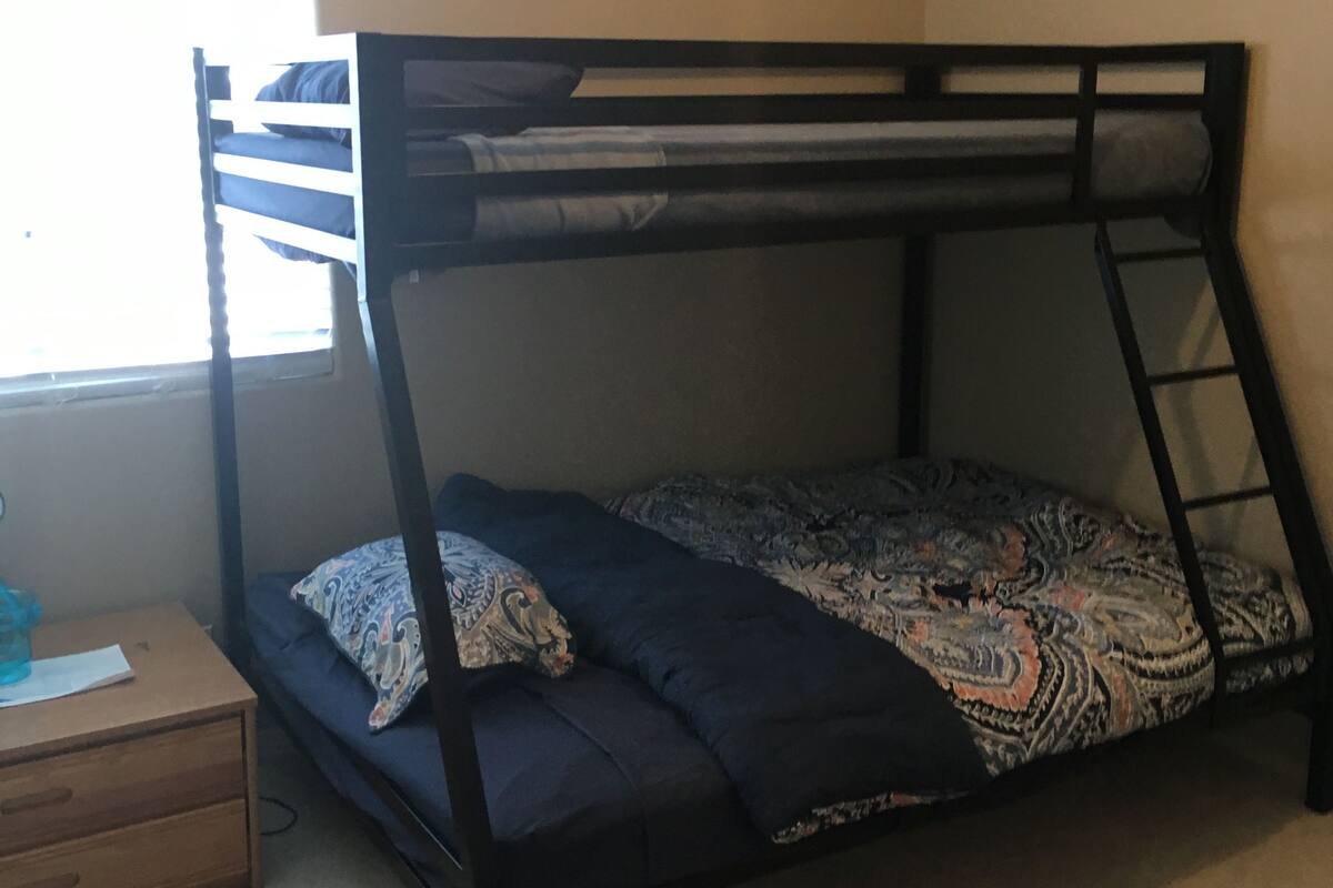 A bunk bed with a ladder, a small wooden nightstand, and a window with blinds.