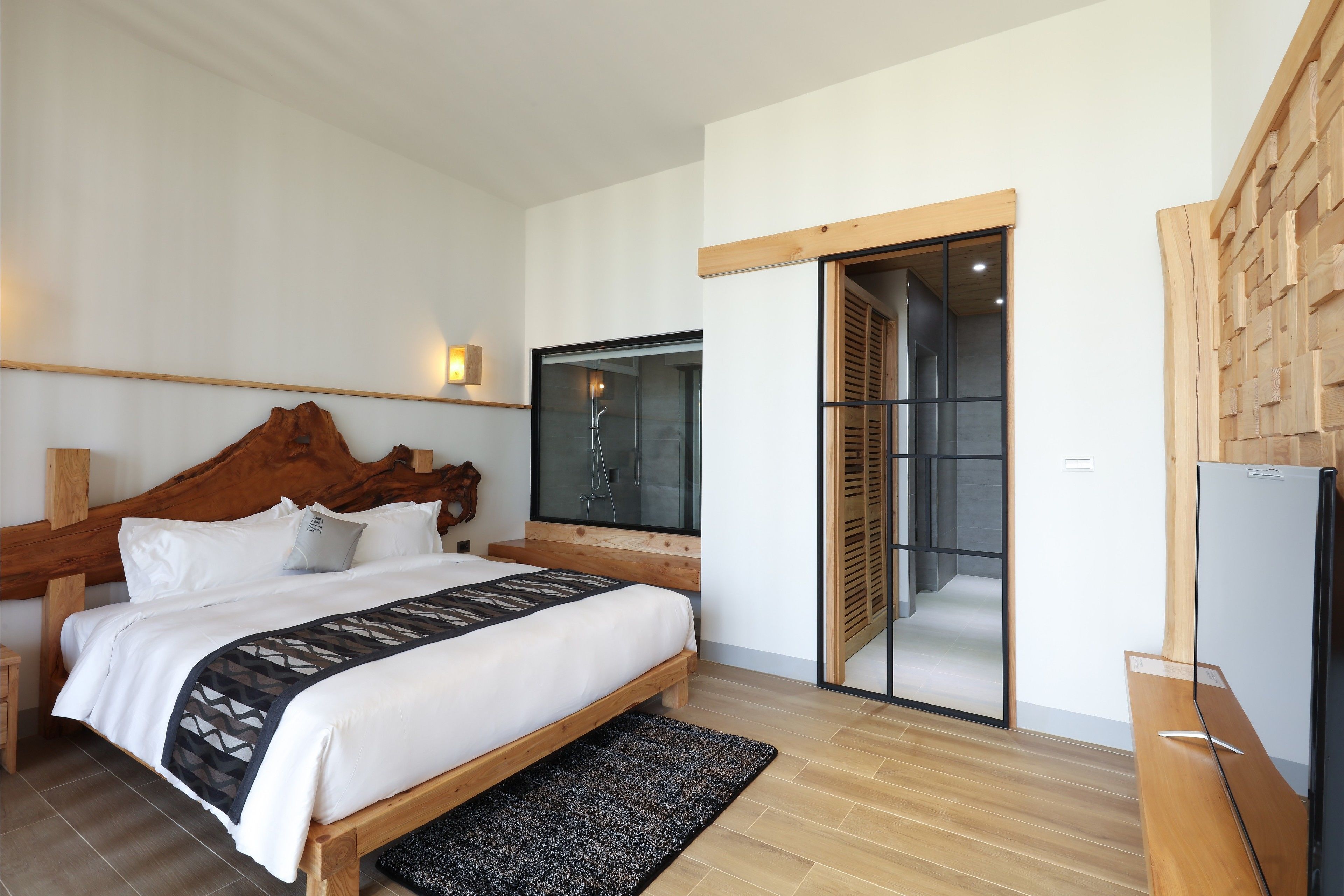 A modern bedroom with a wooden bed, a large window, and a sliding door leading to a shower area.