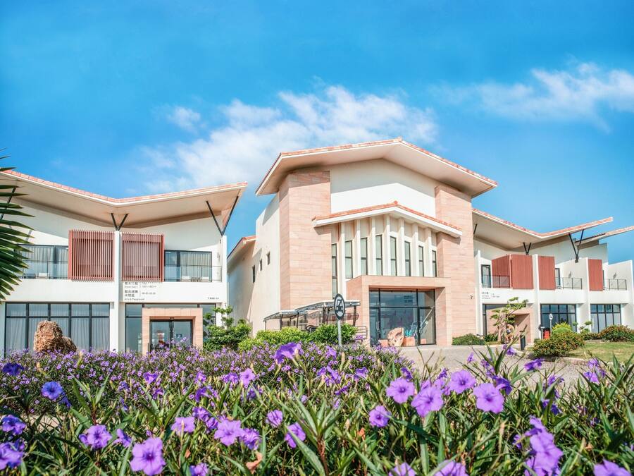 A modern building with a flat roof and large windows, surrounded by a garden of purple flowers.