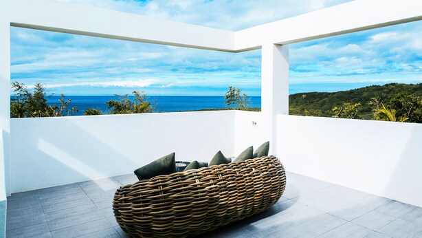 A modern balcony with a wicker seating area, white walls, and a view of the ocean and greenery. - SkyeBay Club (Hengchun)