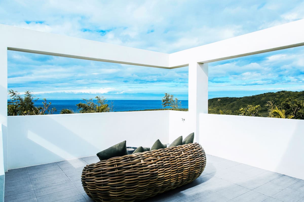 Ein moderner Balkon mit einer Korbsitzgruppe, weißen Wänden und Blick auf das Meer und viel Grün.