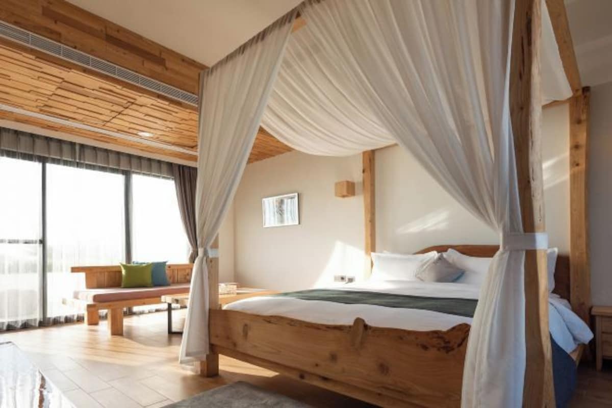 Una cama con dosel de madera con cortinas blancas, un banco y un ventanal con cortinajes.