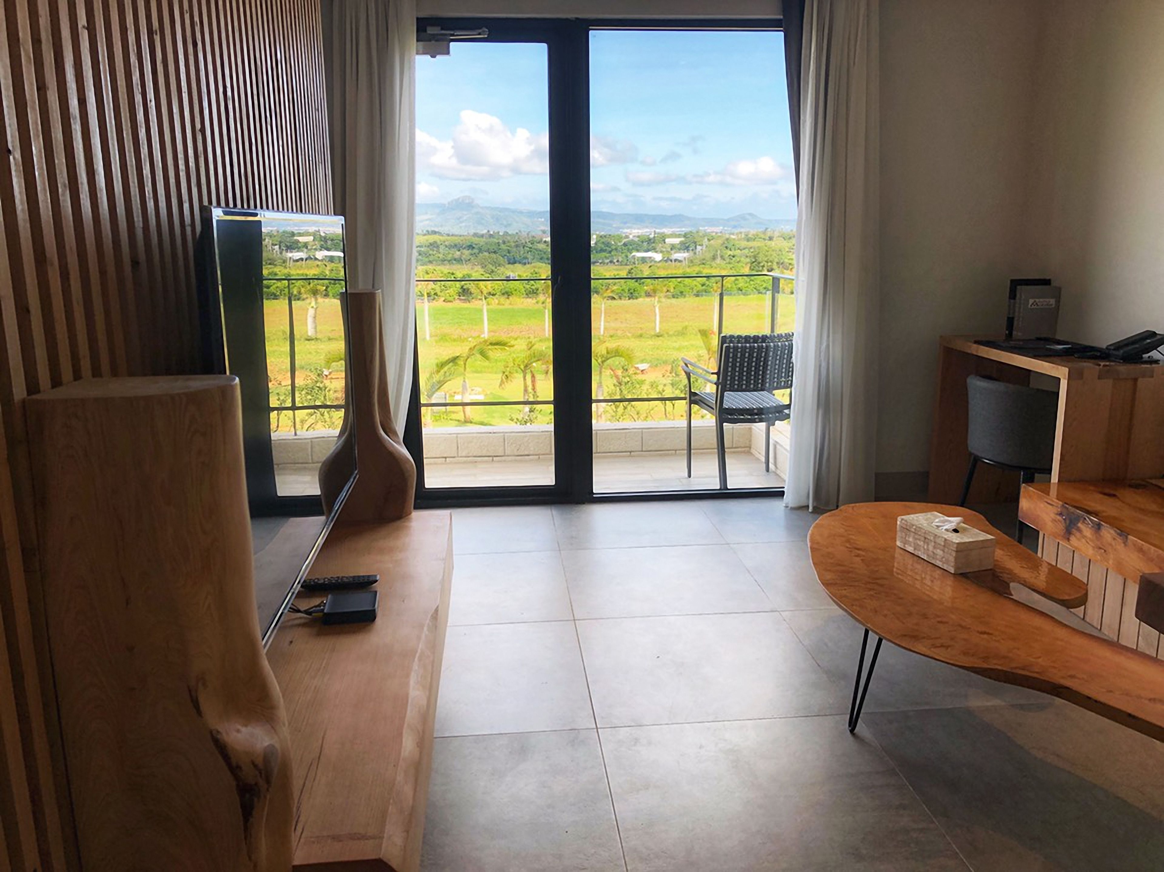 Habitación con banca de madera, una mesita, televisión y balcón con vistas a zonas verdes y montañas.