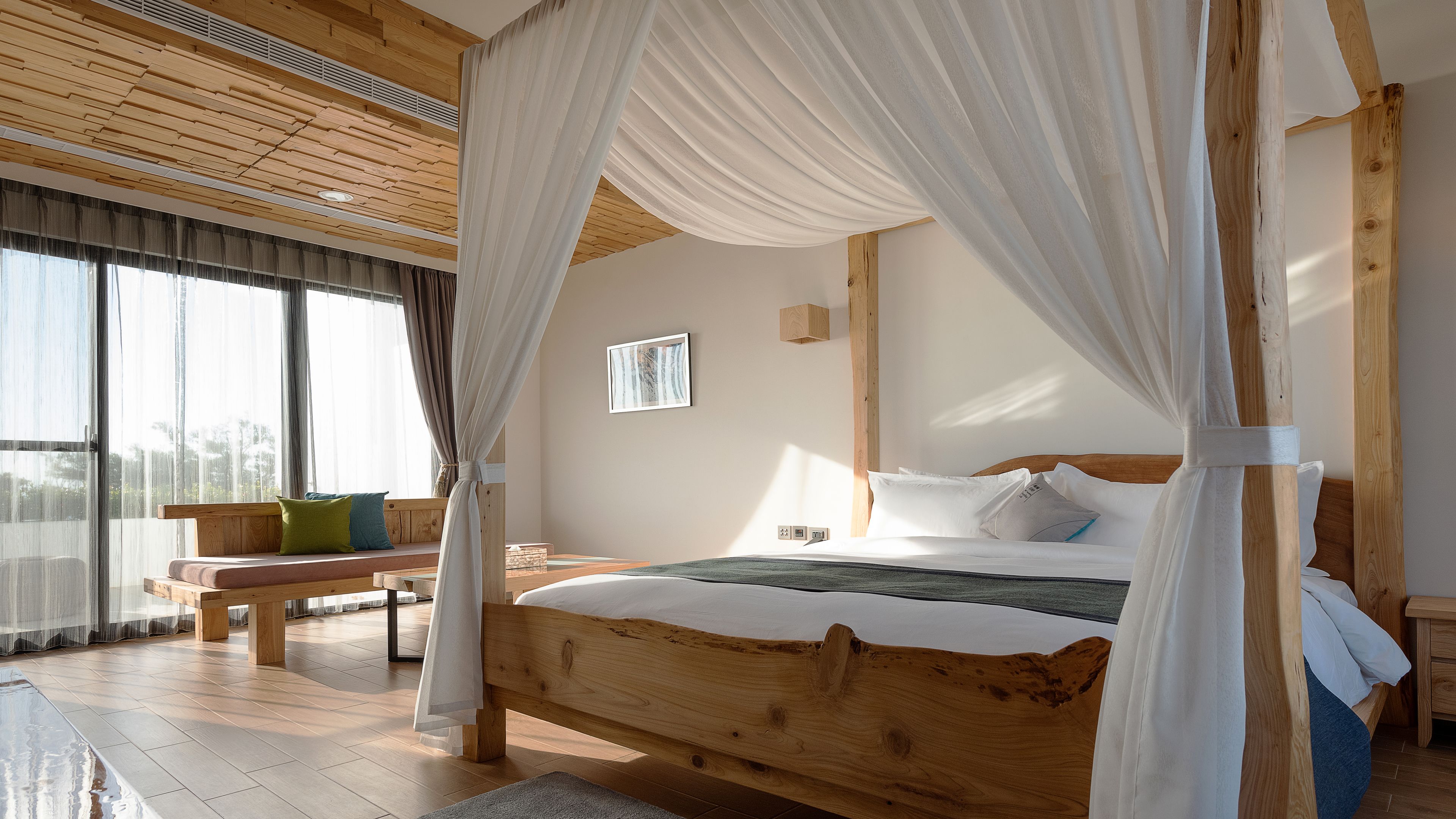 Un dormitorio con cama de madera, un banco y un amplio ventanal con cortinas de gasa.