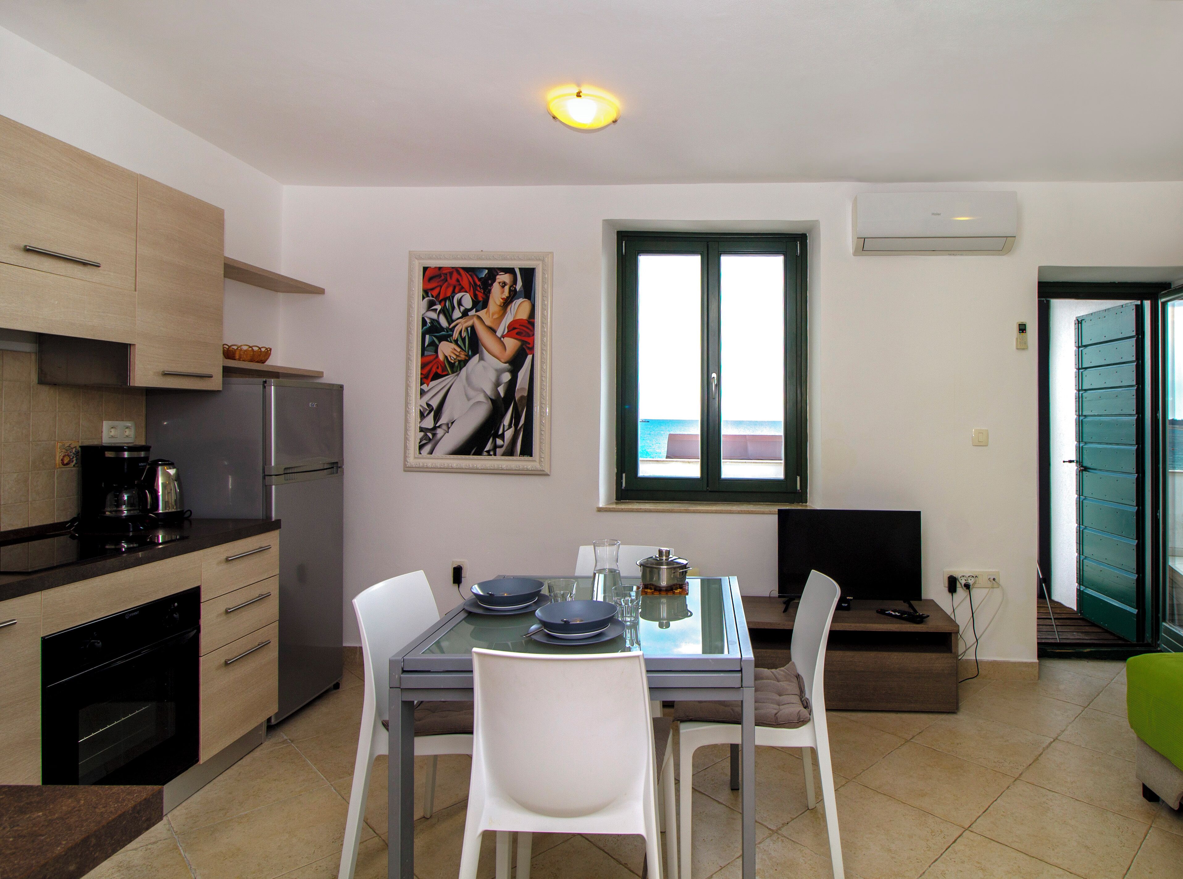 A compact kitchen with modern appliances, a dining table with chairs, a painting on the wall, and a window with a view.