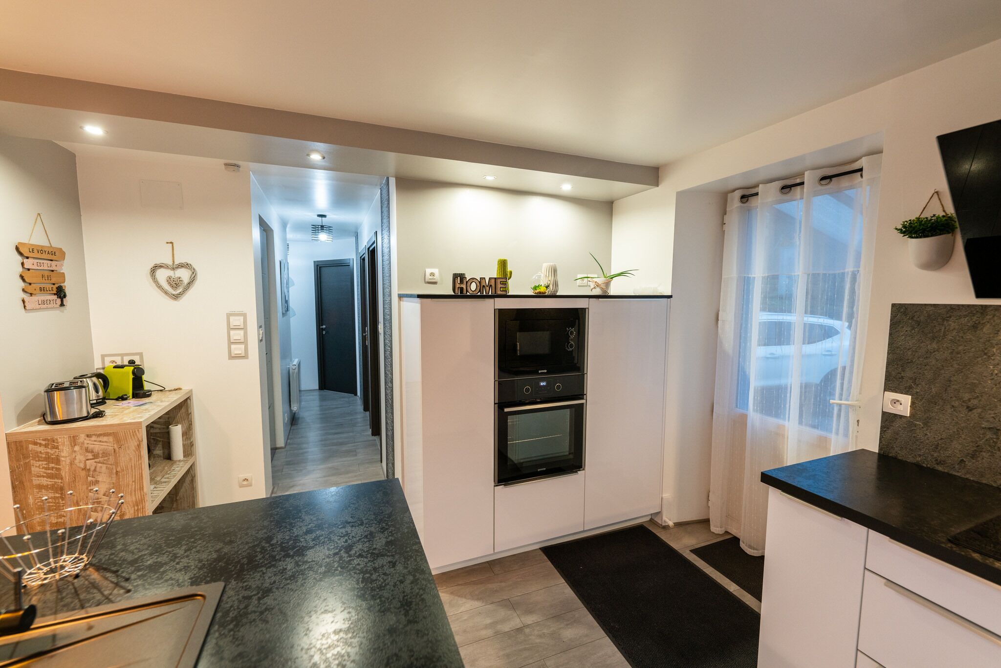 Een moderne keuken met witte kastjes, een zwart aanrecht en een inbouwoven. Er is een houten plank met keukenapparatuur en een hartvormige decoratie.
