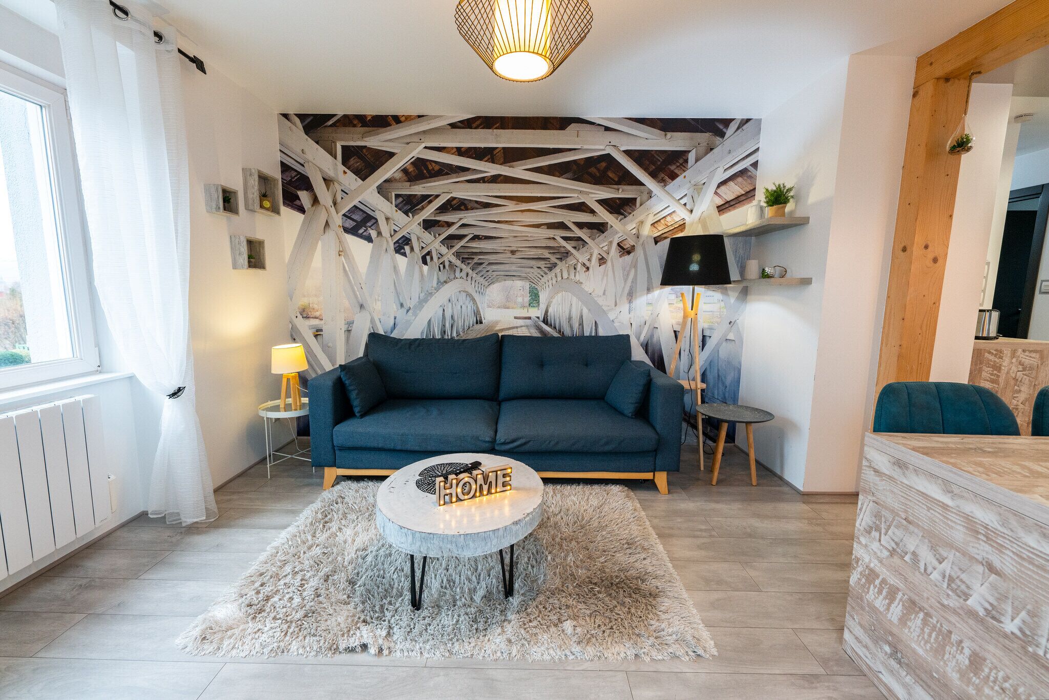 A modern living room with a blue sofa, a round coffee table with a 'HOME' sign, a white fluffy rug, and a wooden ceiling structure.