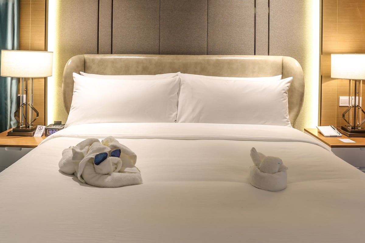Een hotelkamer met een groot bed, twee kussens, een hoofdbord met verticale lijnen, een nachtkastje met een lamp en een wand met een gestructureerd paneel.