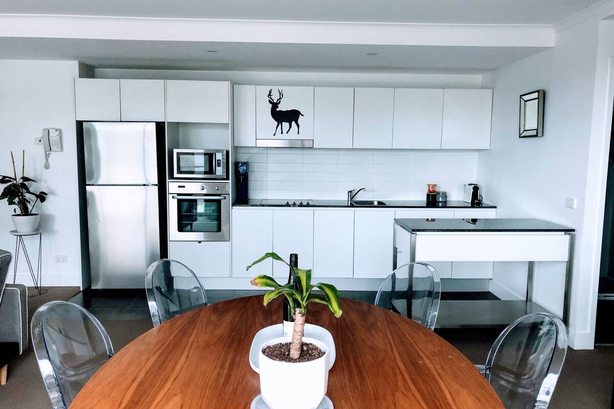 Eine moderne Küche mit weißen Schränken, einem Edelstahlkühlschrank, einem Backofen, einer Mikrowelle und einem Spülbecken. Ein Esstisch mit Stühlen und ein Blumentopf befinden sich in der Mitte.