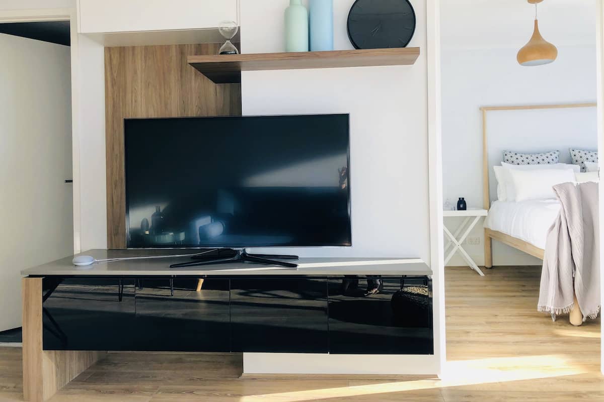 Ein modernes Wohnzimmer mit Flachbildfernseher, Holzschränken, einem Regal mit Dekoration und einem Bett mit gemusterten Kissen.