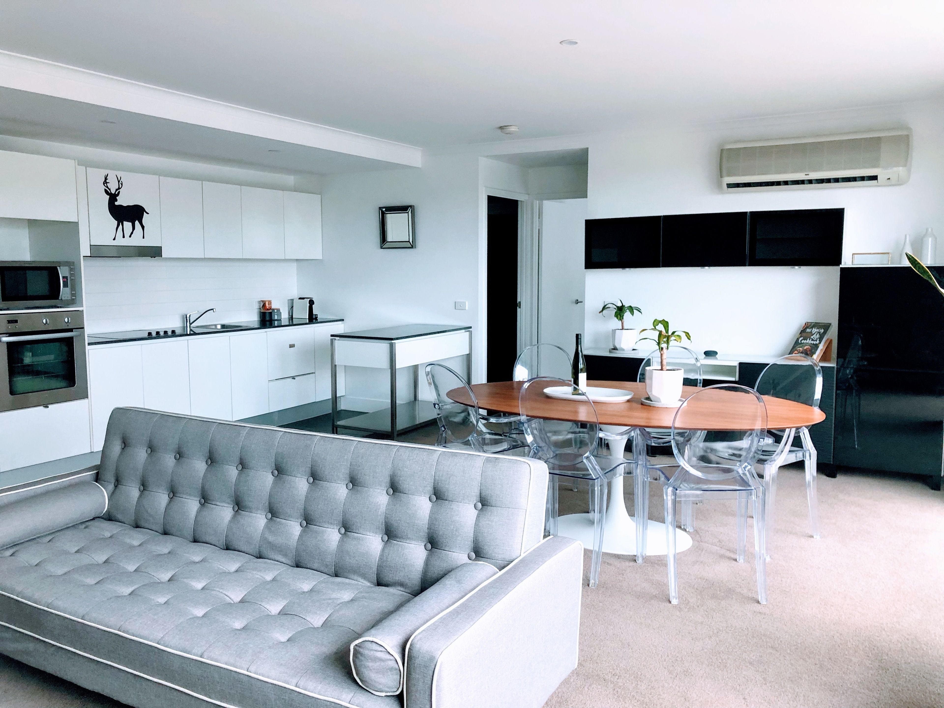 A modern kitchen with white cabinets, a built-in oven, and a microwave. There is a dining area with a wooden table and clear chairs. A grey sofa is in the foreground.