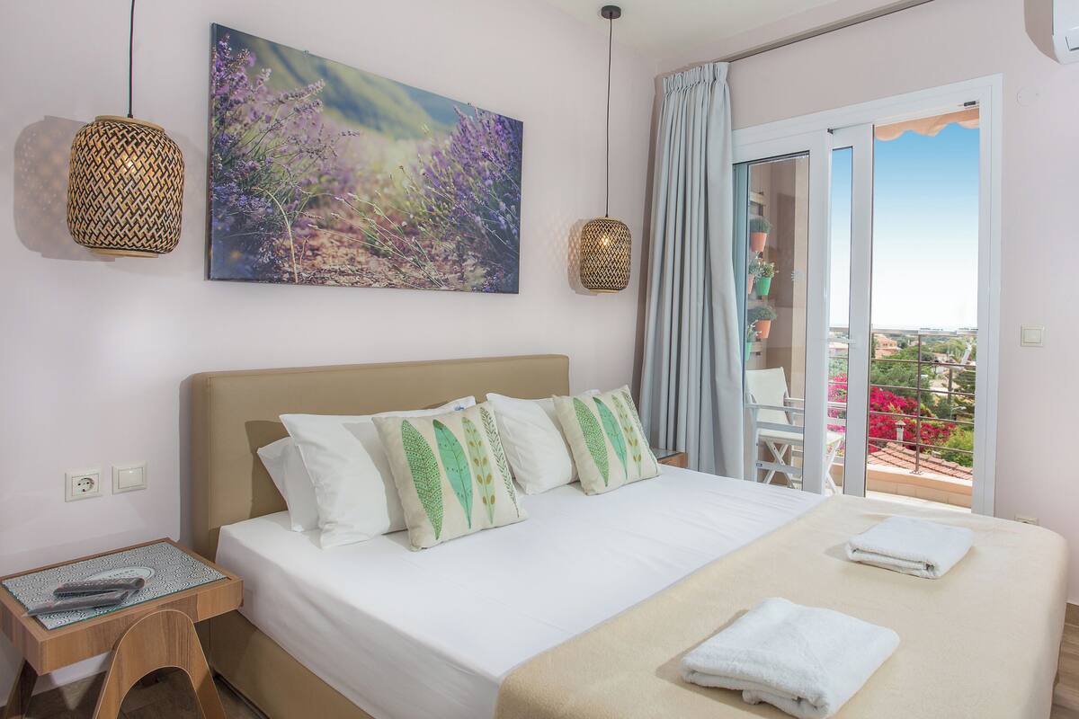 A bedroom with a bed, pillows, a bedside table, a window with curtains, and a balcony with a chair.