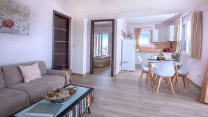 Un salon moderne avec un canapé, une table basse sur laquelle se trouve un panier, et une bibliothèque. On y trouve également un coin repas avec une table et des chaises, et une cuisine visible à travers une porte ouverte.