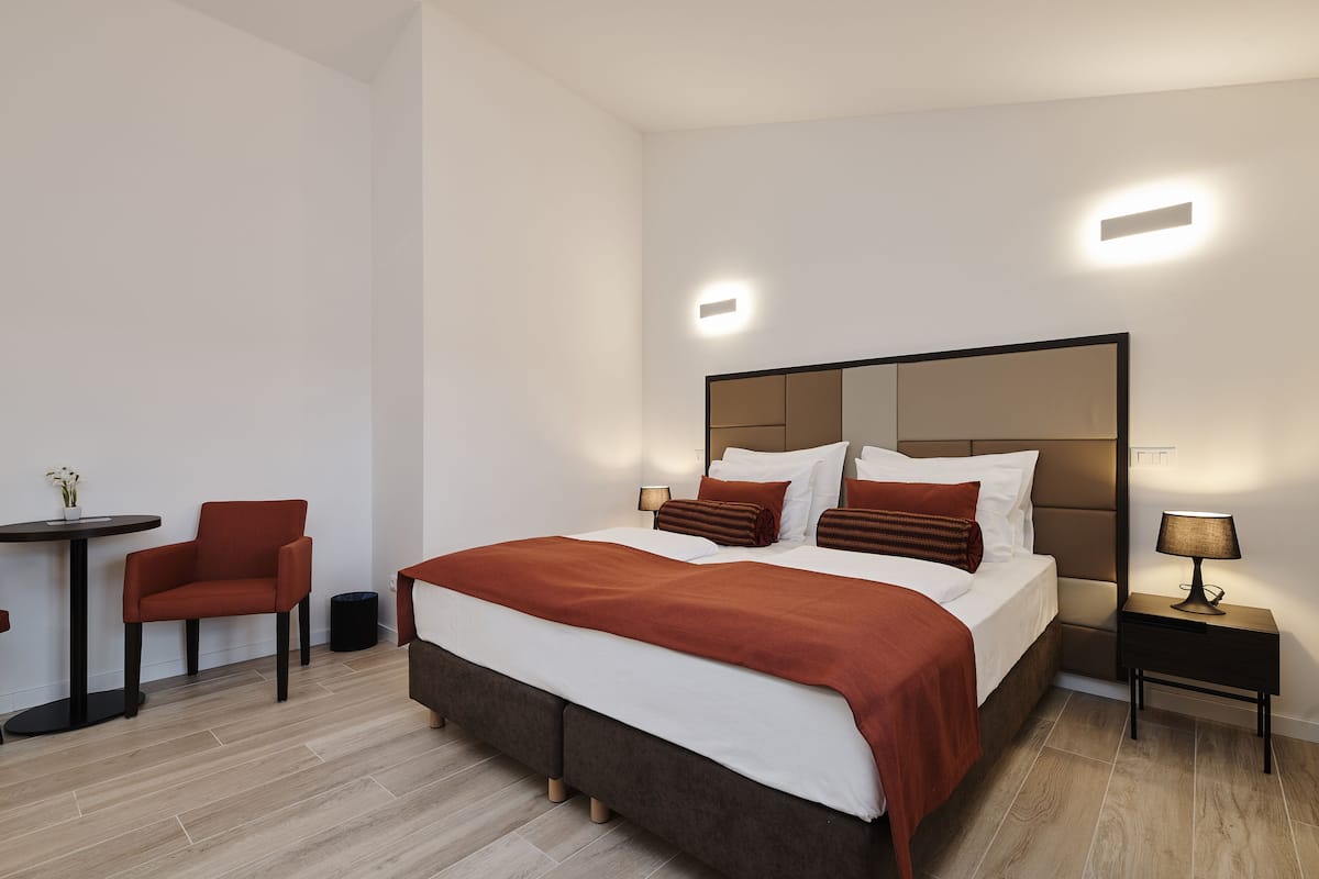 A modern hotel room with a large bed, a bedside table with a lamp, a small round table with a chair, and a potted plant.