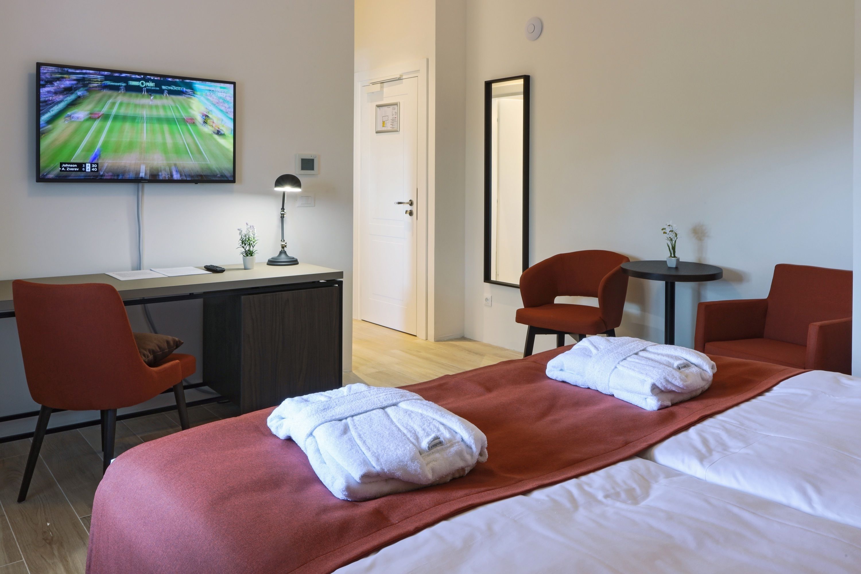A hotel room with a bed, two red chairs, a desk with a TV, and a small table with a plant.
