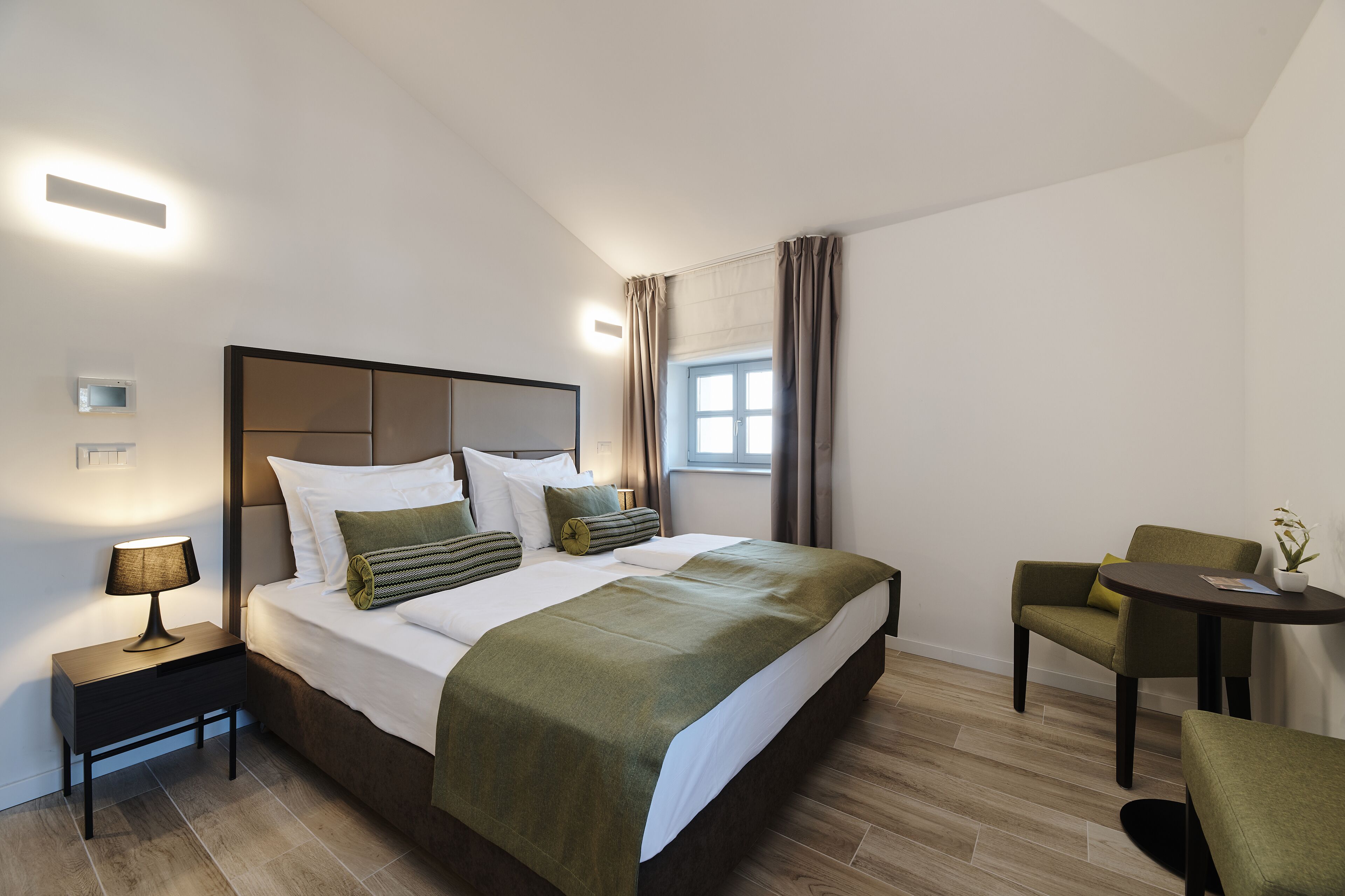 Una habitación de hotel moderna con una cama grande, una mesita de noche con lámpara, una pequeña mesa con planta y una silla.