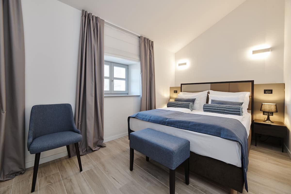 A bedroom with a bed, a chair, a bench, a nightstand, and a window with curtains.