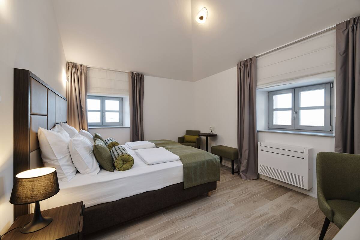 A modern hotel room with a bed, bedside table, lamp, and a view of the city through large windows.