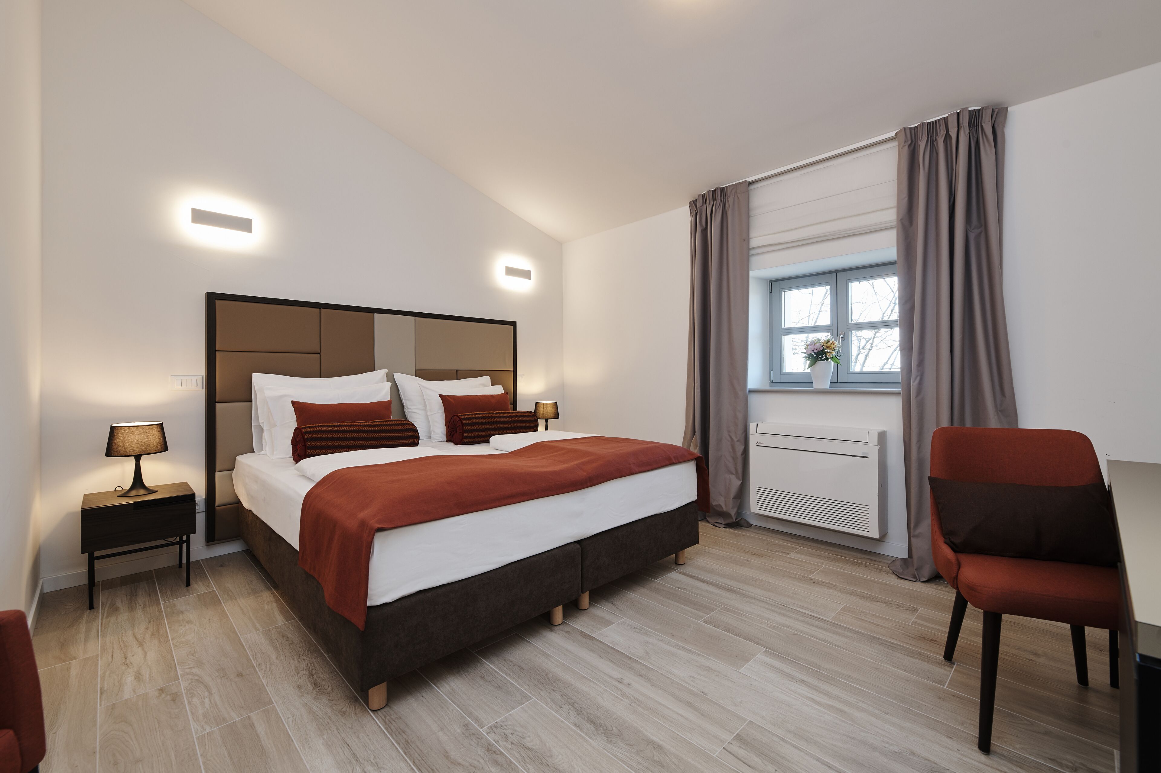 A modern hotel room with a large bed, bedside lamps, a window with curtains, a radiator, and a chair.