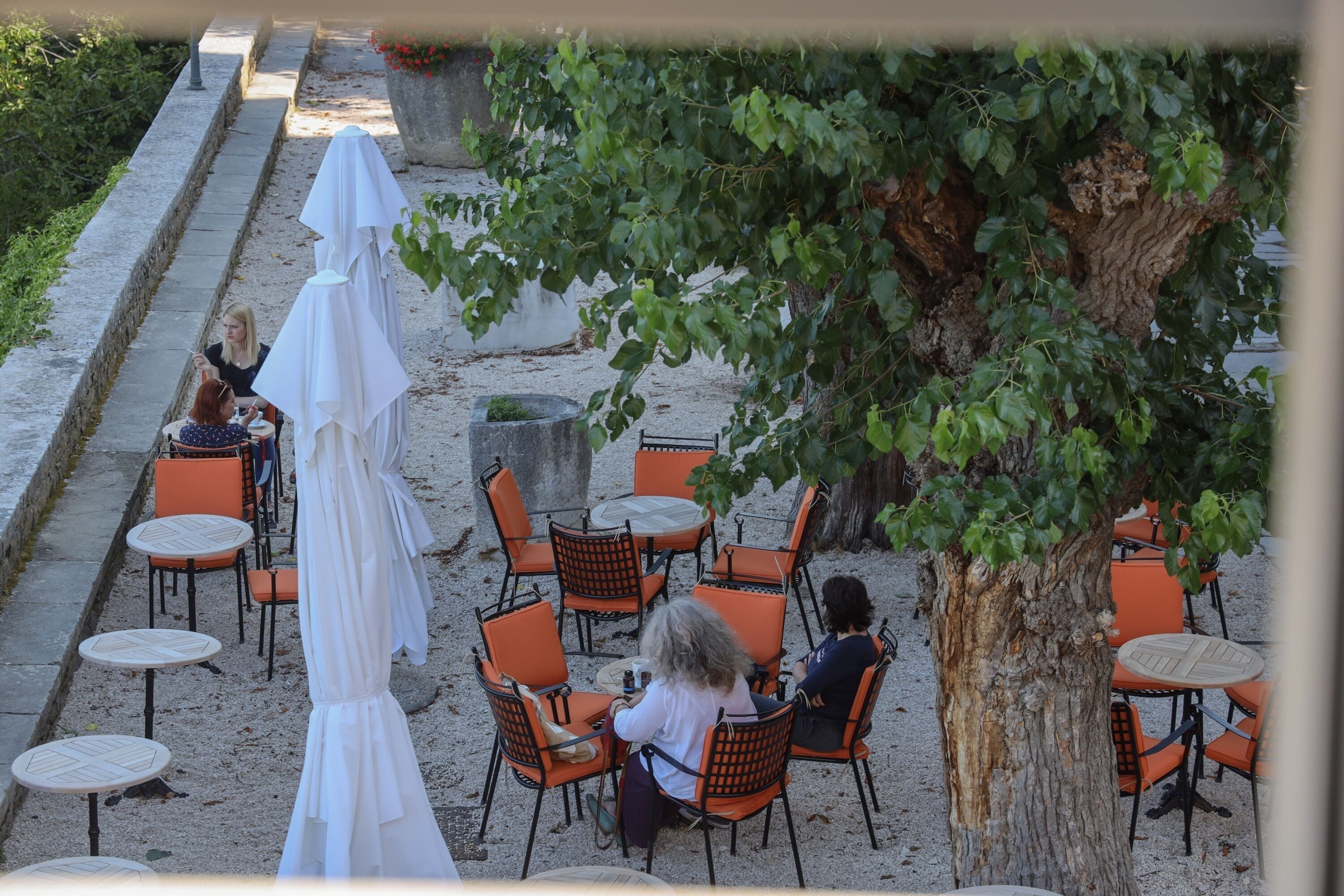 An outdoor seating area with round tables and orange chairs, surrounded by a gravel path and a tree.
