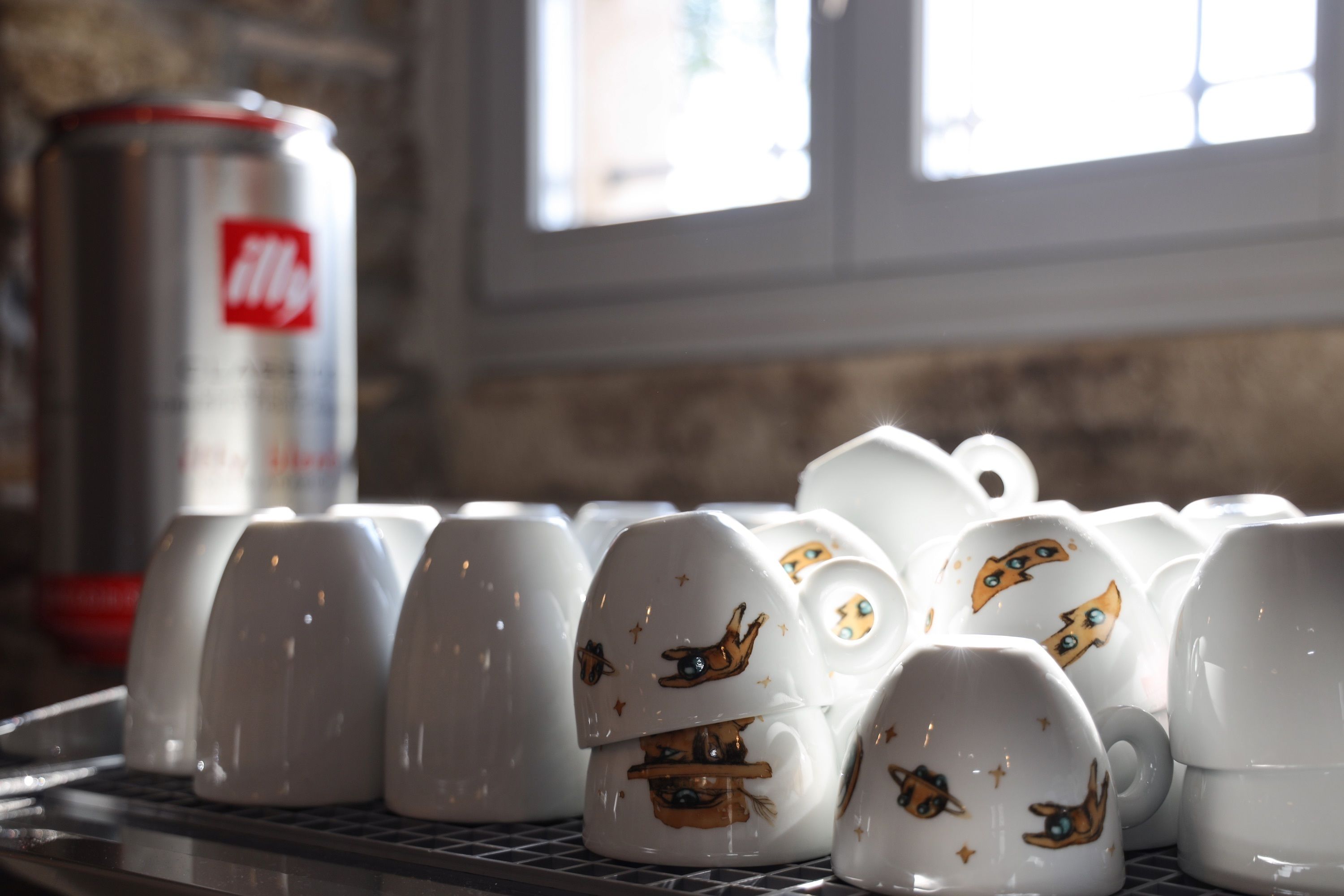 Una hilera de tazas blancas con diseños de gatos sobre un estante metálico, con una lata de Fanta al fondo.