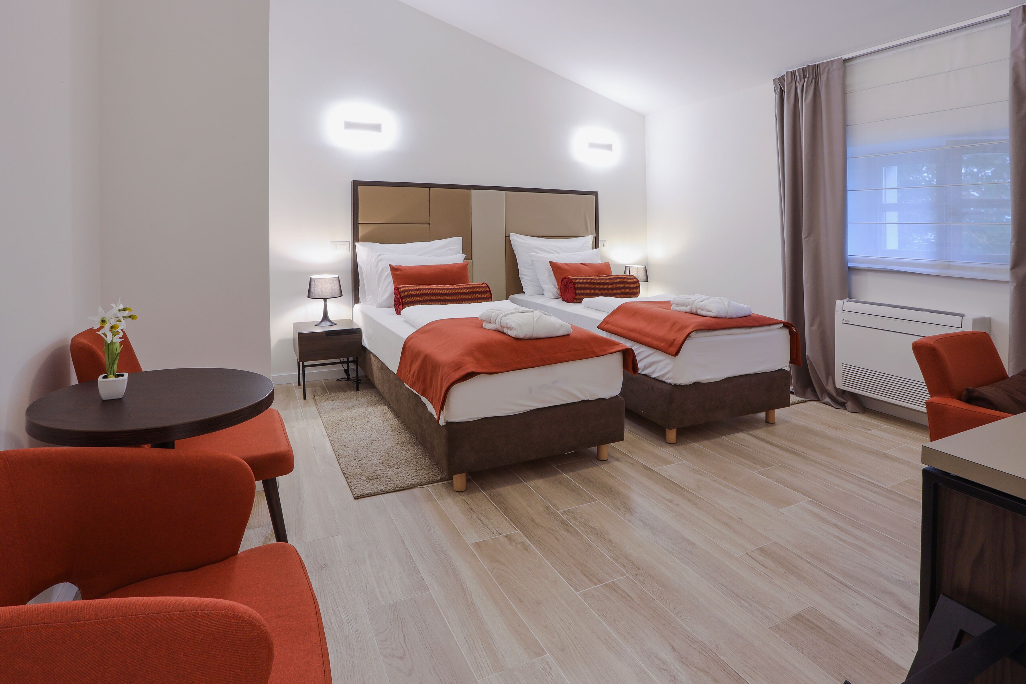 Habitación de hotel con dos camas, una mesita pequeña con una silla, una planta en maceta y una ventana con cortinas.