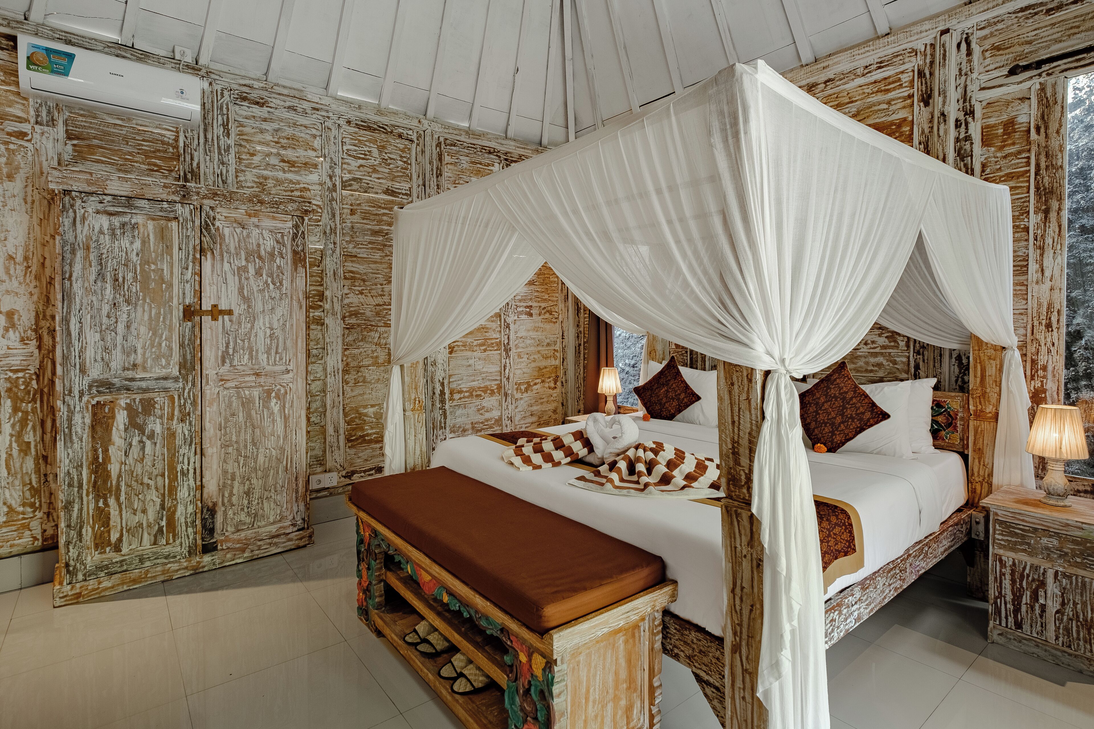 A four-poster canopy bed with a bench, a bedside table with a lamp, and a view of a natural landscape through a window.