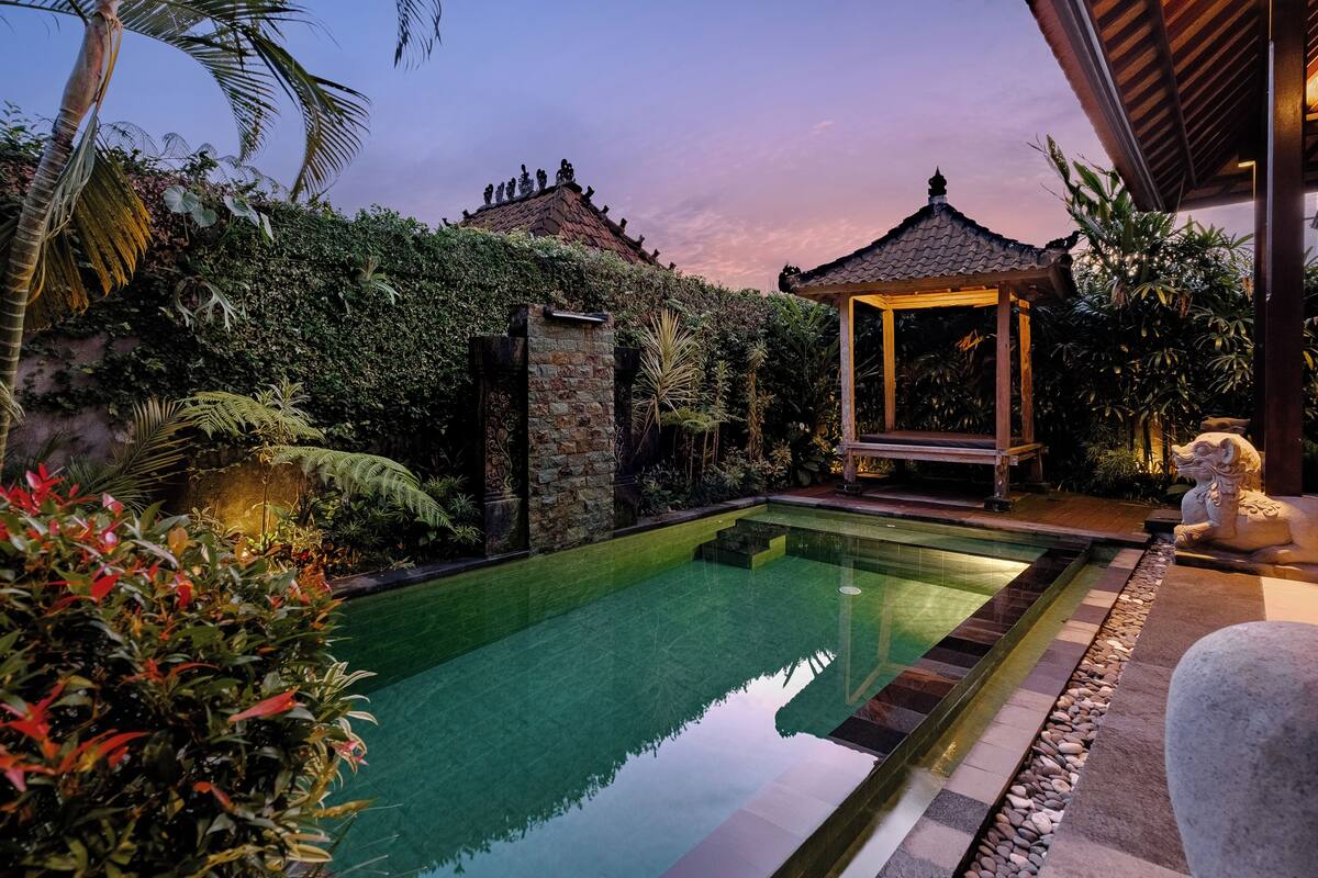 A serene pool area with a stone wall, lush greenery, and a traditional gazebo.