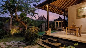 Un espacio exterior bien iluminado con un sendero de piedra, una mesa de madera con sillas, una estatua de piedra y una piscina.
