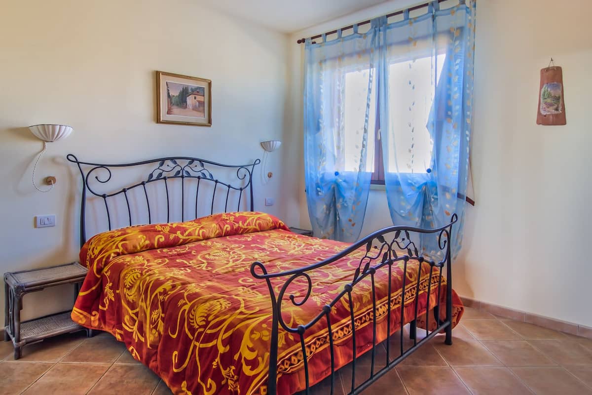 A room with a metal bed frame, a red and yellow patterned bedspread, a small bedside table, a wall-mounted lamp, and a window with blue curtains.