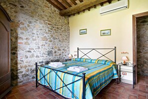 Un dormitorio con pared de piedra, una cama con colcha a rayas, dos cuadros en la pared y una mesita de noche con lámpara.