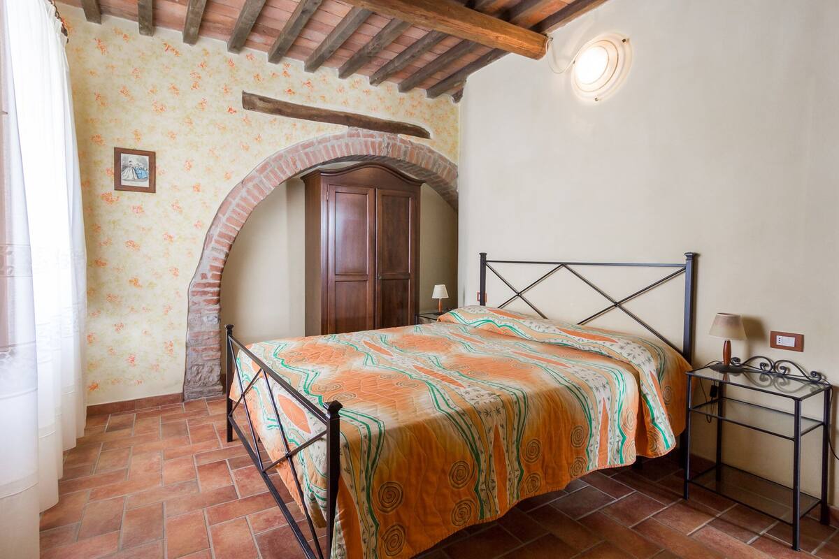 A bedroom with a bed, a nightstand, a lamp, a wooden wardrobe, and a tiled floor.