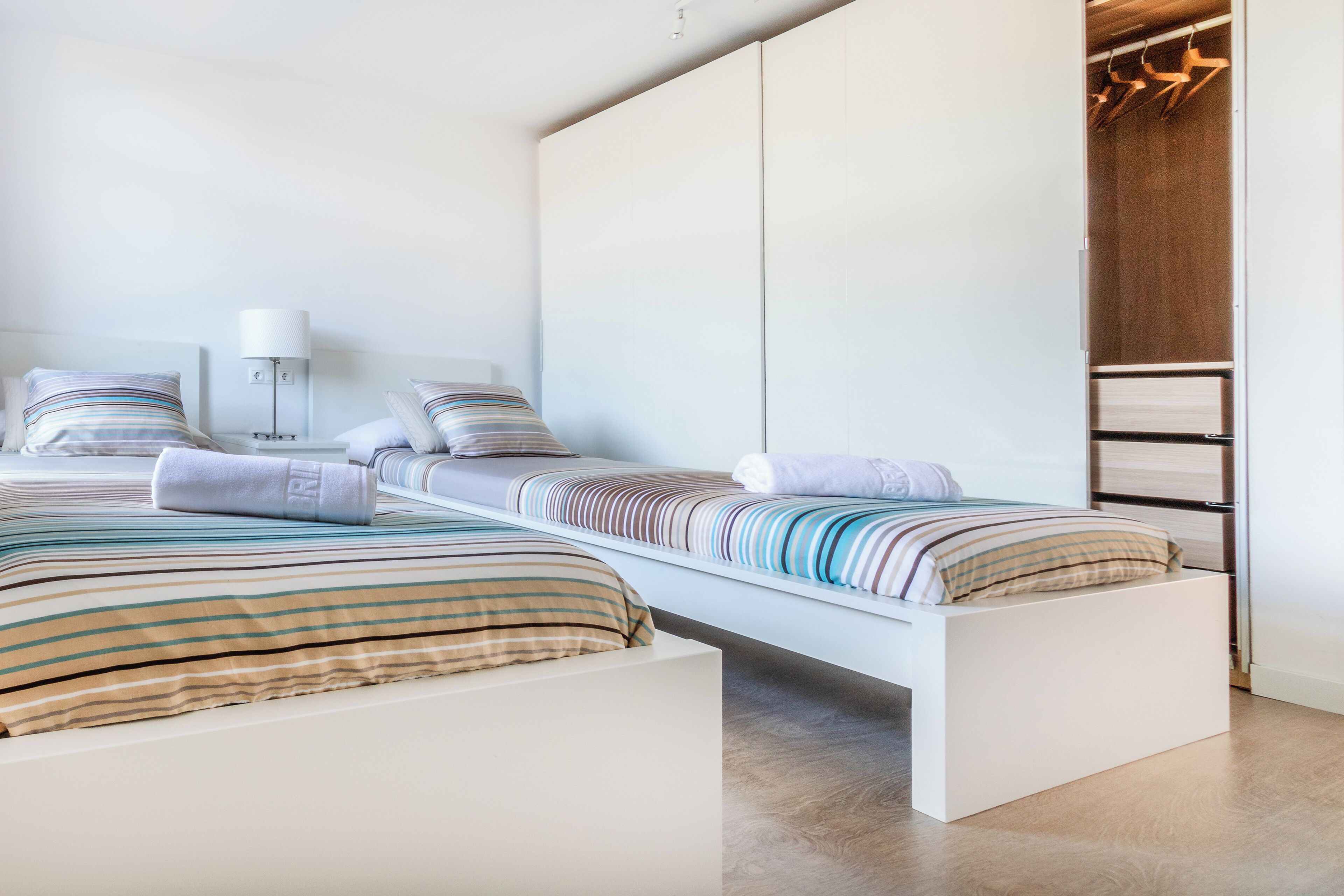 A modern bedroom with two beds, a bedside table with a lamp, and a wooden wardrobe.