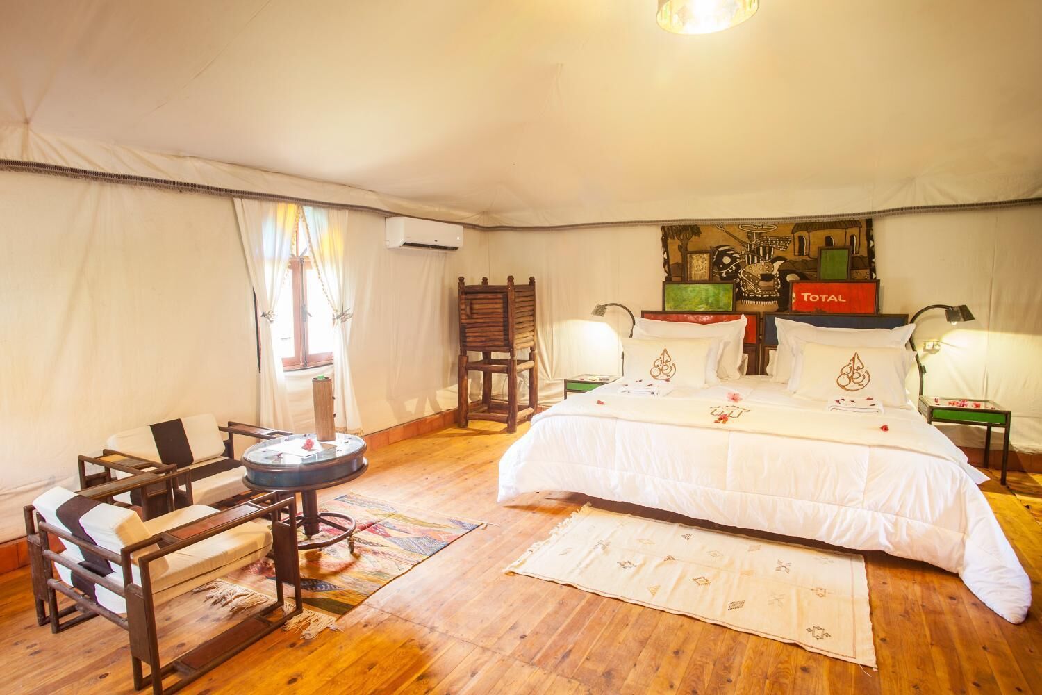 A cozy, well-lit tent-style room with a large bed, a wooden chair, a small table, and a wall-mounted air conditioner.