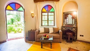 A cozy living room with a sofa, a coffee table, and a decorative mirror. There are two windows with colorful stained glass, and a small wooden stool. - Riad Lamane (Zagora)